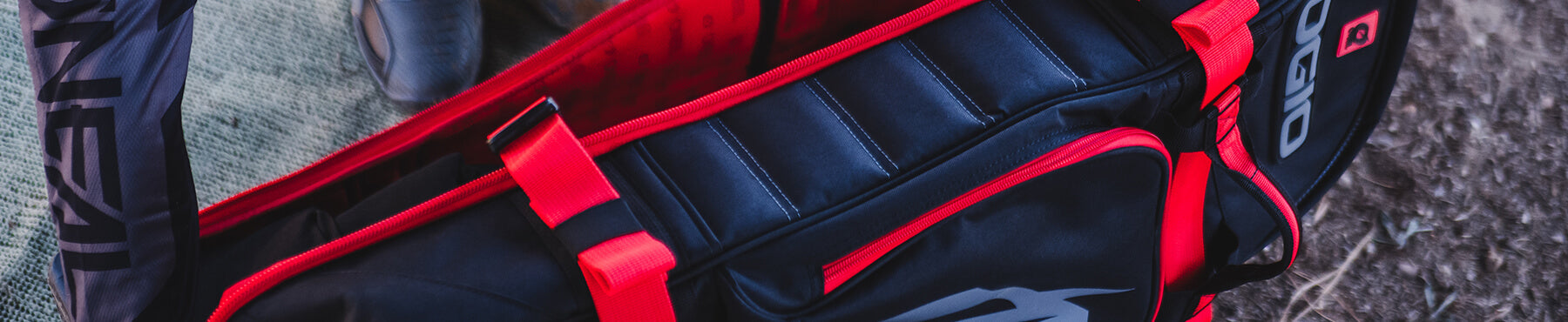 Nahaufnahme einer schwarz-roten Sporttasche mit gepolsterten Abschnitten, Logos und auffälligen Riemen. Die Tasche steht im Freien auf einer strukturierten Oberfläche.