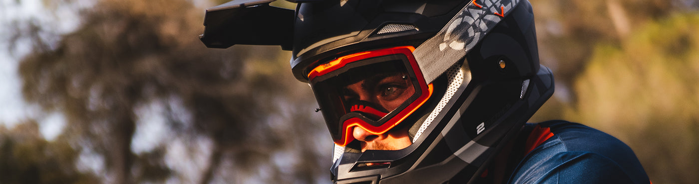 Eine Person, die einen schwarzen Helm mit Visier und eine rote Schutzbrille trägt, befindet sich im Freien, möglicherweise auf einem Mountainbike oder Motorrad, wobei die Bäume im Hintergrund verschwimmen.