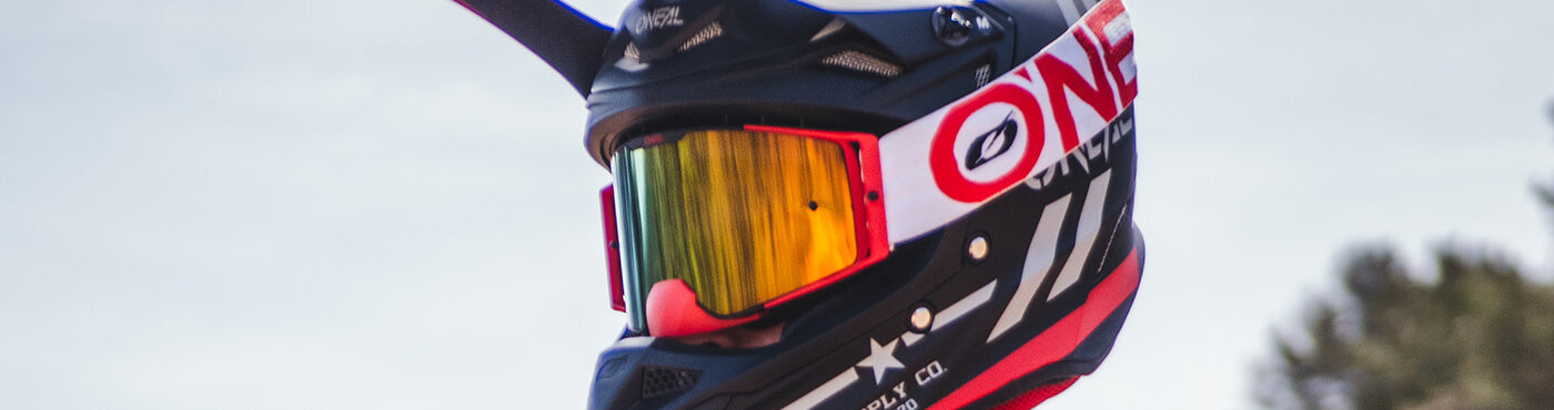 Person, die einen schwarz-roten Motocross-Helm mit reflektierendem Visier trägt, im Freien vor einem hellen Himmel und verschwommenen Bäumen im Hintergrund.