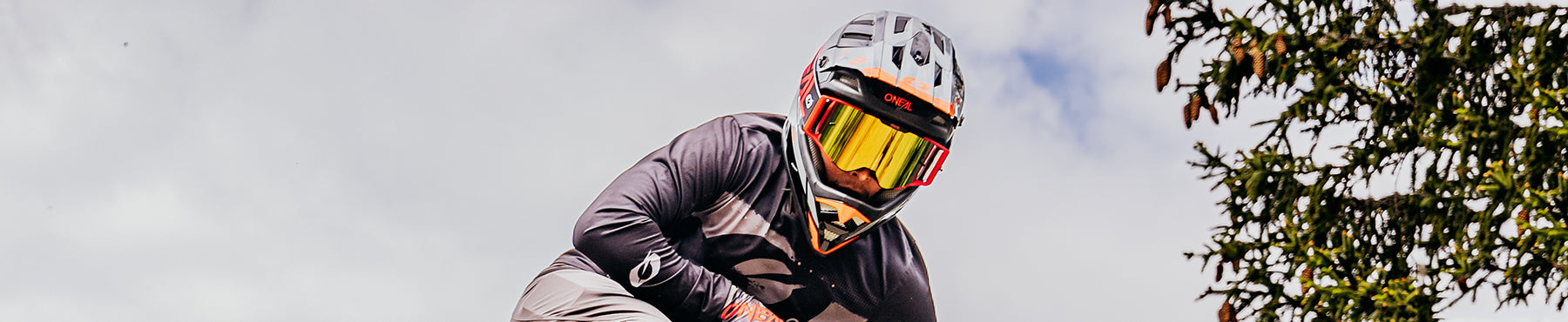 Eine Person mit Integralhelm und Schutzbrille fährt vor einem bewölkten Himmel mit dem Fahrrad bergab. Auf der rechten Seite sind grüne Blätter zu sehen.