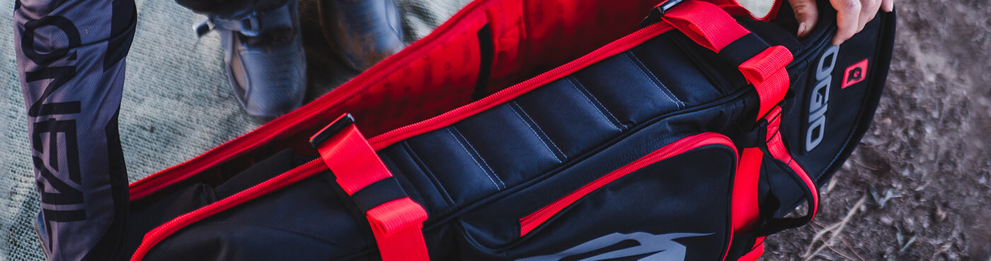 Eine Person in dunkler Sportkleidung trägt eine schwarz-rote Sporttasche mit gepolstertem Riemen und steht auf einer Oberfläche aus Erde und Netzgewebe.
