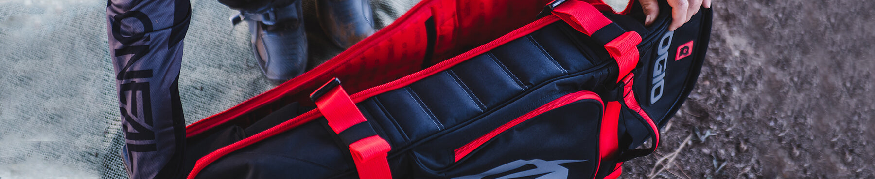 Eine Person in dunkler Sportkleidung trägt eine schwarz-rote Sporttasche mit gepolstertem Riemen und steht auf einer Oberfläche aus Erde und Netzgewebe.