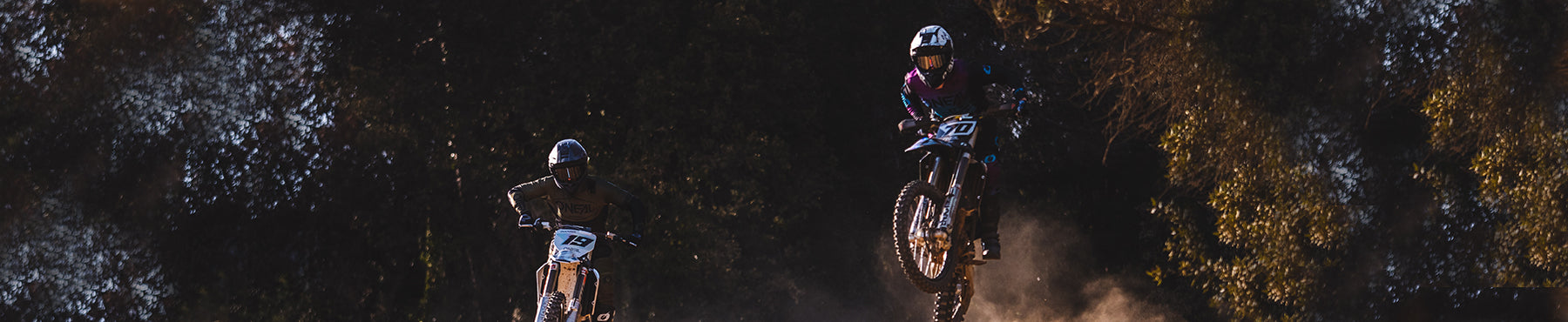 Zwei Motocrossfahrer in der Luft vor einer Baumkulisse. Ein Fahrer links mit weißem Motorrad, Helm und dunkler Ausrüstung; der Fahrer rechts trägt ein buntes Outfit, beide wirbeln Staub auf.