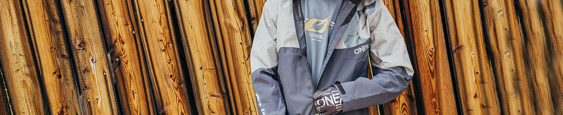 Person, die eine grau-blaue O'Neal-Jacke mit passenden Handschuhen trägt, steht vor einem Holzzaun und hält ihre Hände zusammen.