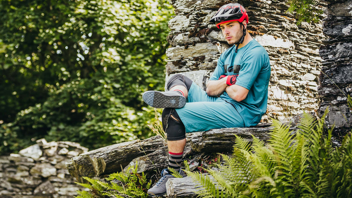 Eine Person mit rotem Helm und blauer Radlerkleidung sitzt auf einer Steinkonstruktion und verschränkt Arme und Beine. Sie trägt Knie- und Ellbogenschützer und ist von üppigen grünen Farnen und Blättern umgeben.