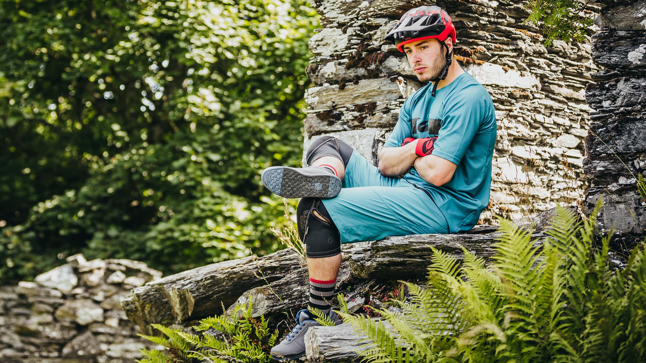 Eine Person mit rotem Helm und blauer Radlerkleidung sitzt auf einer Steinkonstruktion und verschränkt Arme und Beine. Sie trägt Knie- und Ellbogenschützer und ist von üppigen grünen Farnen und Blättern umgeben.