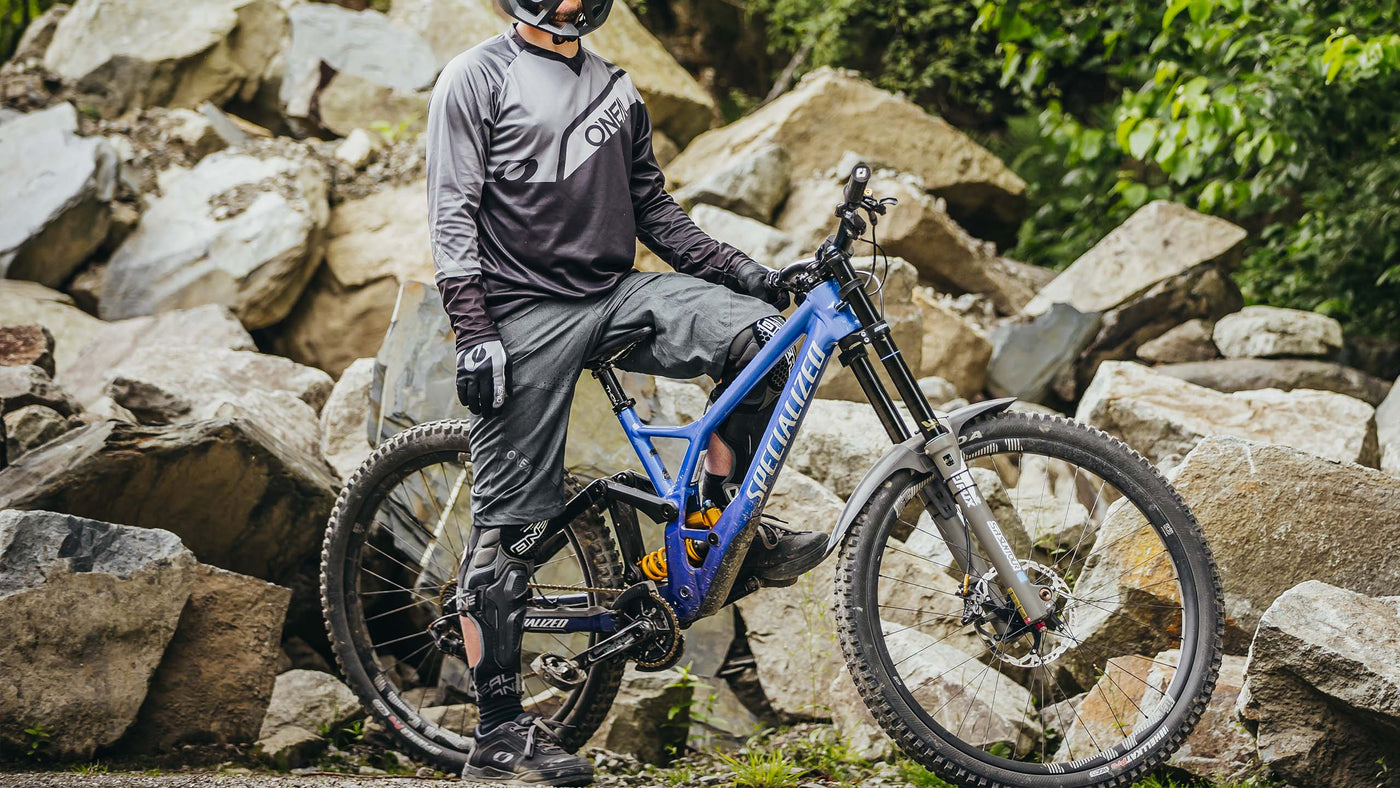 Eine Person in Schutzausrüstung steht mit einem blauen Mountainbike auf einem felsigen Pfad. Sie ist von großen Felsen umgeben und im Hintergrund sieht man Grün.