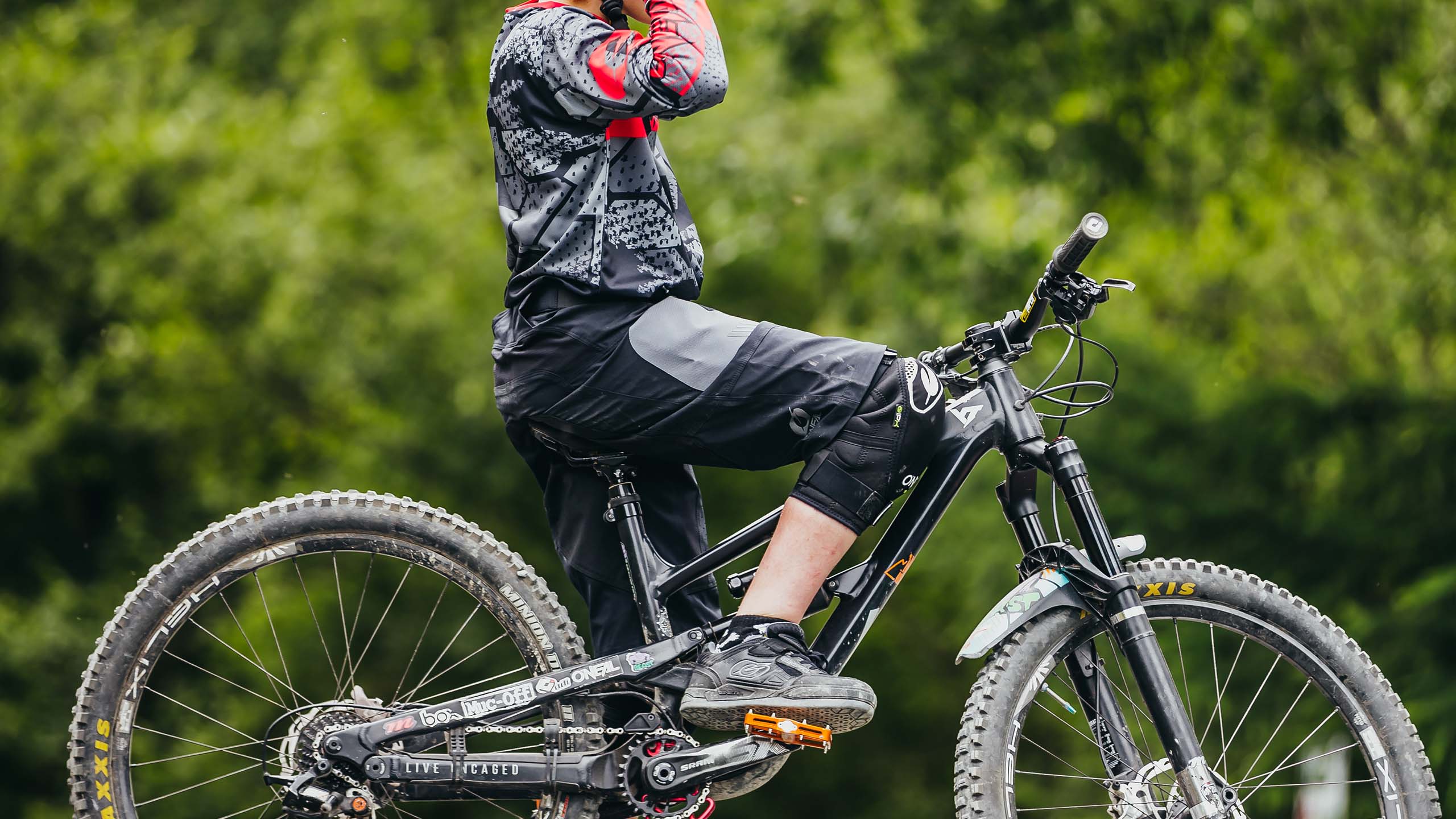 Eine Person in einem rot-schwarz gemusterten Trikot sitzt auf einem Mountainbike und rückt einen Helm zurecht. Das Fahrrad ist schwarz mit dicken Reifen und im Hintergrund ist ein verschwommener grüner Wald zu sehen.