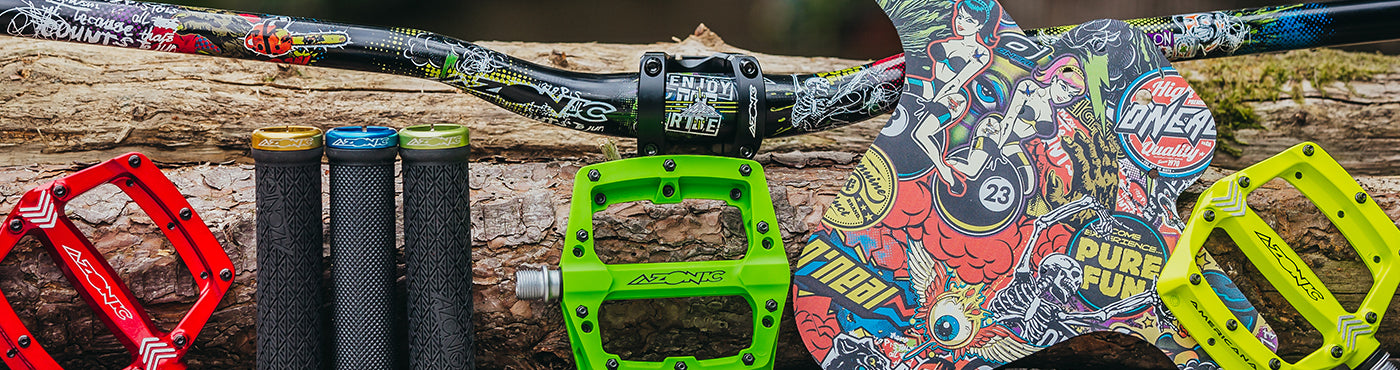 Auf einem Baumstumpf sitzt eine bunte Auswahl an Fahrradkomponenten, darunter Griffe, Pedale und ein Kettenblatt mit lebendigen Designs. Die Komponenten sind im Graffiti-Stil gestaltet und in kräftigen Farben wie Rot, Gelb und Grün gehalten.