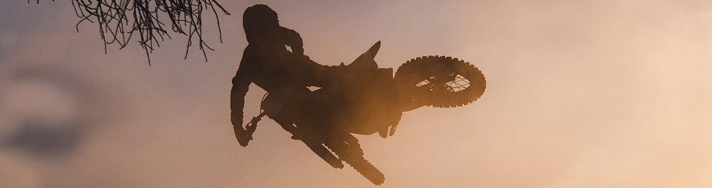 Ein Motocross-Fahrer hebt sich vor einem orangefarbenen Himmel ab und springt mit seinem Motorrad hoch in die Luft, umgeben von Ästen.