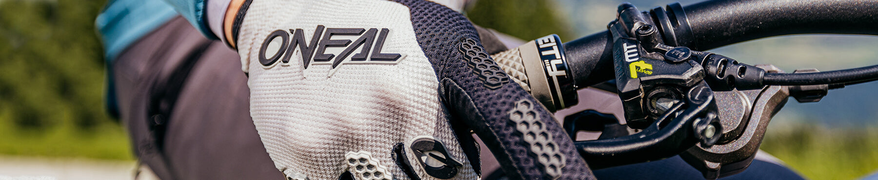 Nahaufnahme der behandschuhten Hand eines Radfahrers, die einen Fahrradlenker umklammert. Auf dem Handschuh ist die Marke „O'Neal“ zu sehen. Im Hintergrund ist verschwommenes Grün zu sehen, was eine Szenerie im Freien suggeriert.