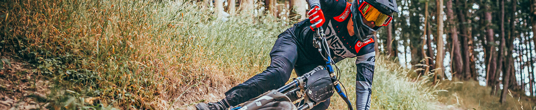 Ein Mountainbiker in Schutzausrüstung lehnt sich in eine Kurve auf einem von Bäumen und Gras umgebenen Feldweg.