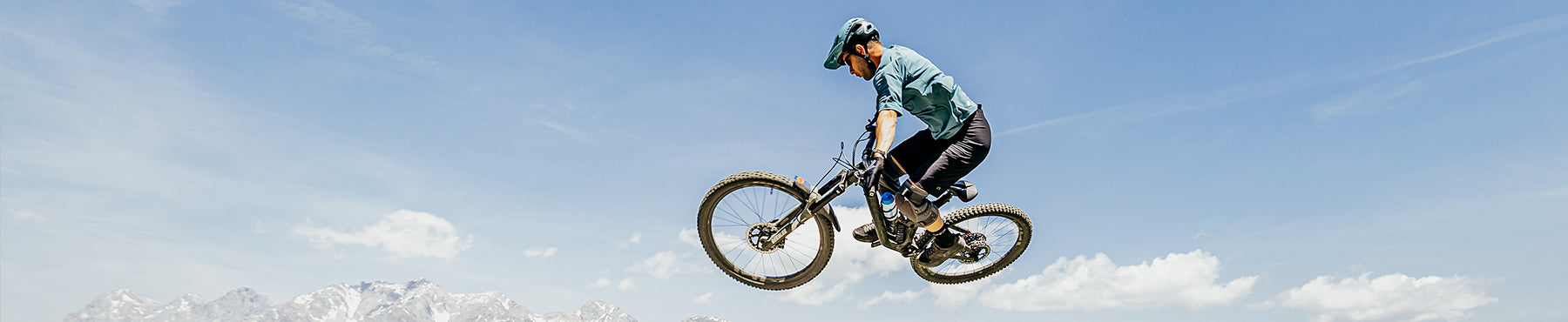 Ein Radfahrer mit Helm und blauem Hemd führt vor der Kulisse eines klaren blauen Himmels und der Berge in der Ferne auf einem Mountainbike einen Stunt in der Luft aus.
