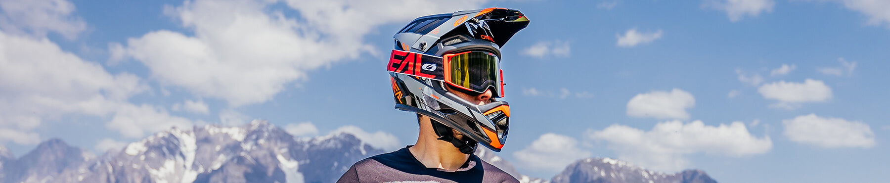 Eine Person mit Motocross-Helm und Schutzbrille steht vor einer Bergkulisse und einem blauen Himmel mit vereinzelten Wolken.