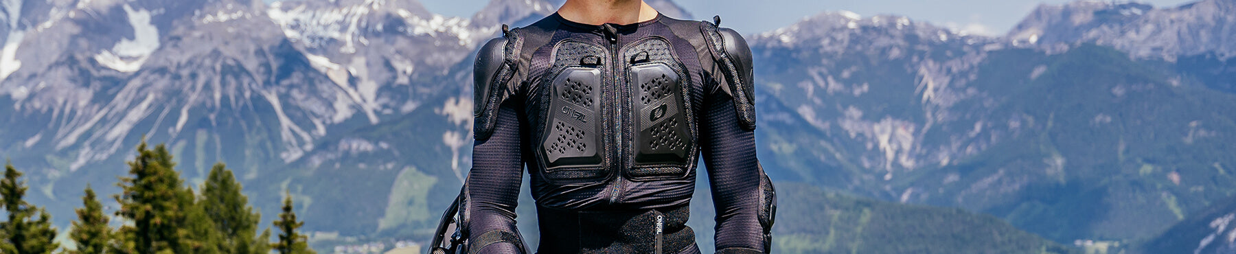 Eine Person in Schutzkleidung steht im Freien vor der Kulisse majestätischer Berge und Kiefern unter einem klaren blauen Himmel.