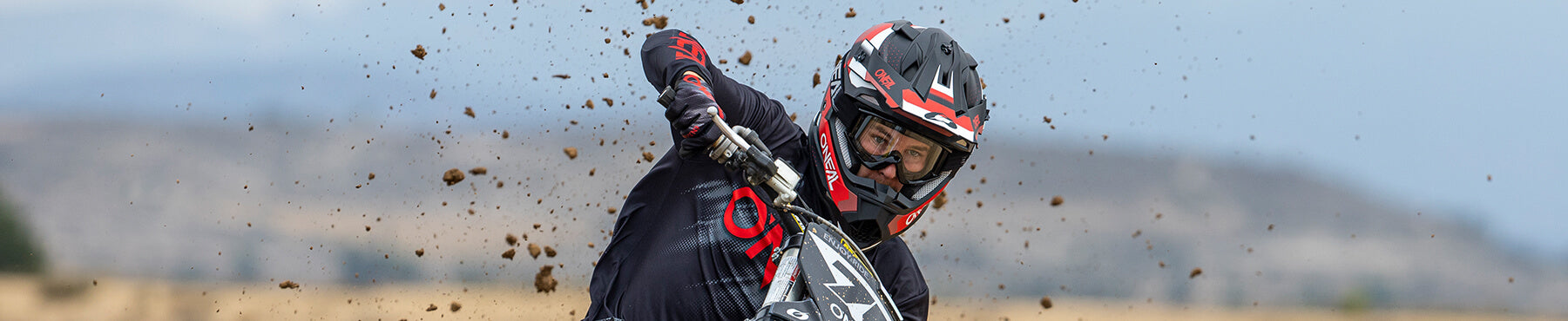 Ein Motocrossfahrer mit Helm und Schutzbrille lehnt sich in eine Kurve und wirbelt dabei Staub über die Strecke. Im Hintergrund ist eine verschwommene Landschaft mit Hügeln unter einem bewölkten Himmel zu sehen.