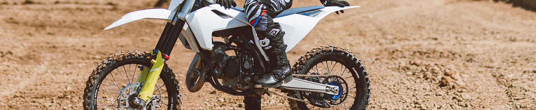 Eine Person in Motocross-Ausrüstung fährt mit einem weißen Dirtbike auf einer unbefestigten Strecke, hinter ihr sind Staubspuren zu sehen. Der Hintergrund ist leicht verschwommen, was die Bewegung und Geschwindigkeit betont.