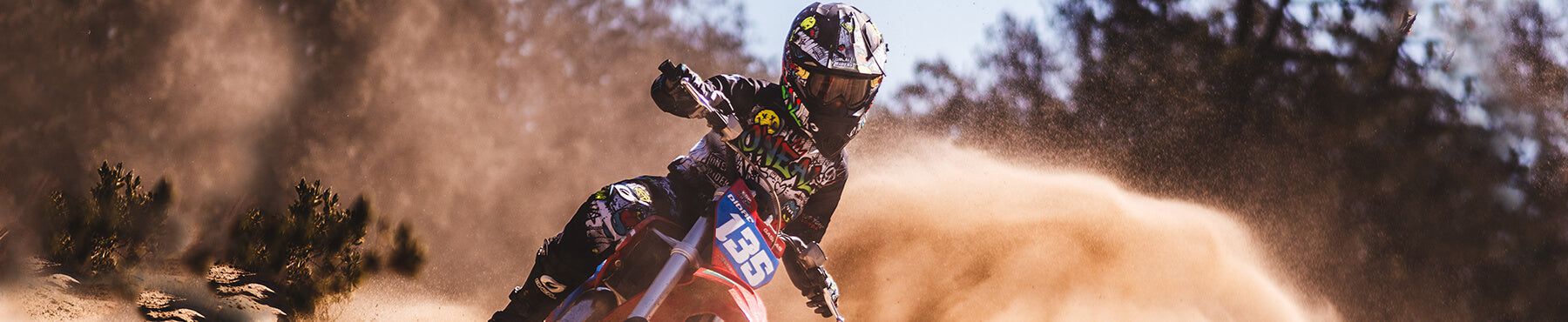 Ein Motocrossfahrer in voller Montur rast über eine Sandpiste, umgeben von Staubwolken. Der Fahrer legt sich in eine Kurve und erzeugt vor dem Hintergrund von Bäumen und blauem Himmel ein dynamisches Gefühl von Bewegung und Intensität.
