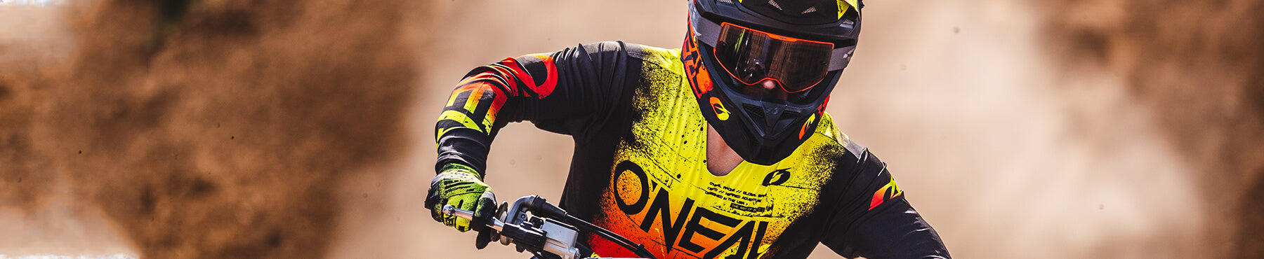 Ein Motocross-Fahrer in farbenfroher Schutzausrüstung und Helm rast umgeben von Staubwolken über eine unbefestigte Strecke.