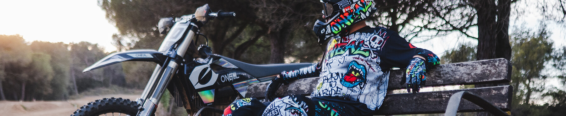 Eine Person in bunter Motocross-Ausrüstung und Helm sitzt auf einer Bank neben einem Dirtbike in einem Waldstück, im Hintergrund geht die Sonne unter.