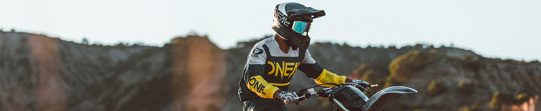 Eine Person in Motocross-Ausrüstung, einschließlich Helm und Schutzbrille, fährt an einem sonnigen Tag ein Dirtbike. Im Hintergrund sind felsige Hügel und ein klarer Himmel zu sehen.