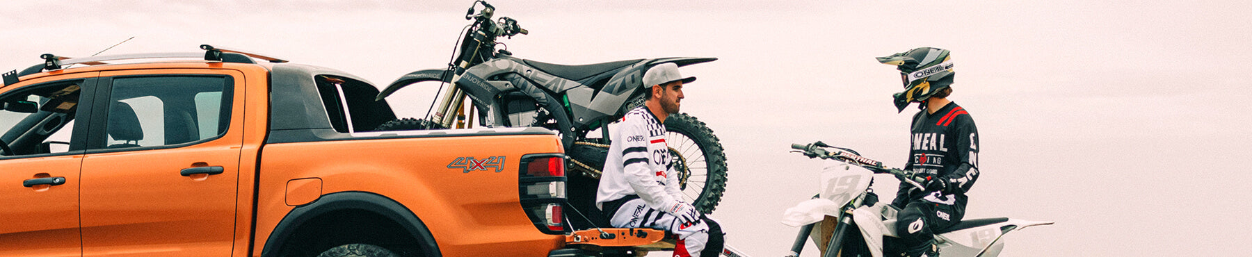Zwei Personen in Motocross-Ausrüstung sitzen auf der Ladeklappe eines orangefarbenen Pickups, auf dem ein schwarzes Dirtbike geladen ist. Eine Person sitzt daneben auf einem weißen Dirtbike. Die Szene spielt sich vor einem bewölkten Himmel ab.