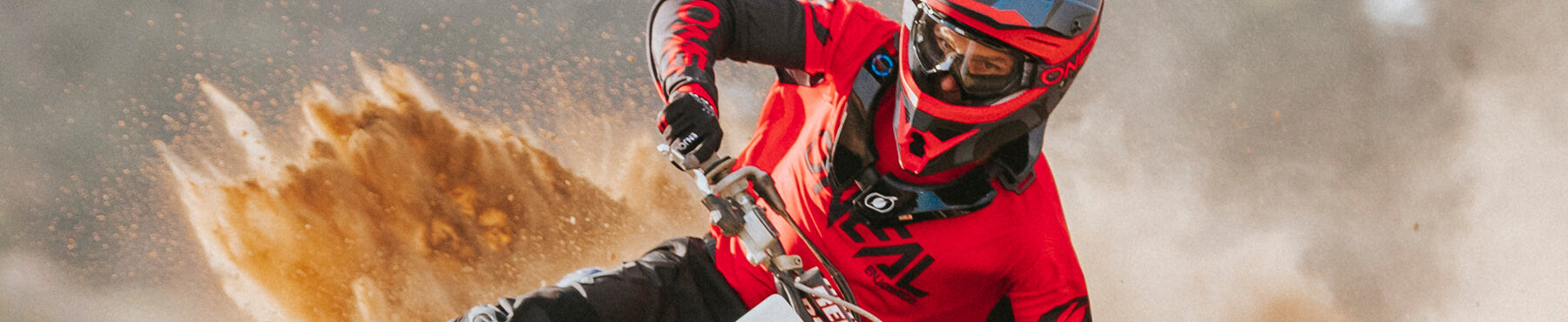 Ein Motocrossfahrer in rot-schwarzem Outfit wirbelt Staub auf, als er auf einer Rennstrecke eine scharfe Kurve fährt. Staubwolken bilden sich um das Motorrad und der Fahrer trägt zum Schutz einen roten Helm mit Schutzbrille.