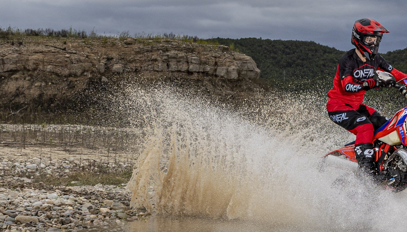 Eine Person in roter und schwarzer Ausrüstung fährt mit einem Dirtbike durch einen seichten Fluss mit spritzendem Wasser vor einer Kulisse aus felsigem Gelände und Bäumen unter einem bewölkten Himmel.