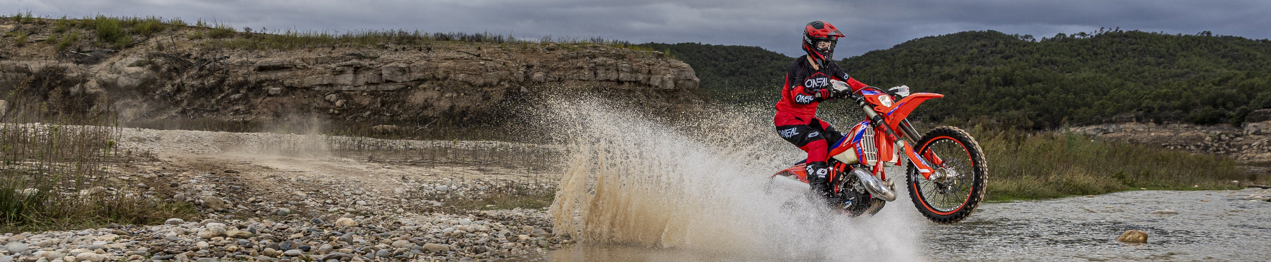 Eine Person in roter und schwarzer Ausrüstung fährt mit einem Dirtbike durch einen seichten Fluss mit spritzendem Wasser vor einer Kulisse aus felsigem Gelände und Bäumen unter einem bewölkten Himmel.