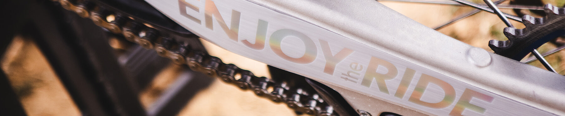 Nahaufnahme eines Fahrradrahmens mit dem Text „Enjoy the Ride“ in Farbverlauf. Das Bild konzentriert sich auf die Kette und einen Teil des Getriebemechanismus, wobei das Sonnenlicht die metallische Oberfläche sanft beleuchtet.