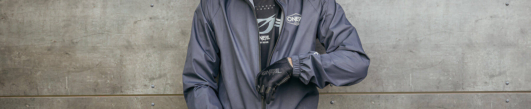 Eine Person in dunkelgrauer Jacke und schwarzen Handschuhen richtet ihren Ärmel. Im Hintergrund ist eine strukturierte Betonwand zu sehen. Nur der Oberkörper ist zu sehen.