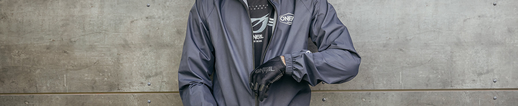 Eine Person in einer dunkelgrauen Jacke und Handschuhen steht vor einer Betonwand. Der Reißverschluss der Jacke ist teilweise geöffnet, so dass ein Logo auf dem Hemd sichtbar ist. Das Foto ist am Hals und an der Taille abgeschnitten.