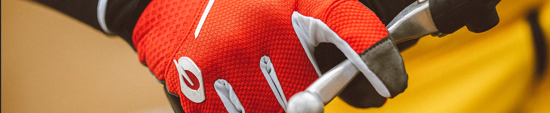 Nahaufnahme einer Person, die rot-weiße strukturierte Handschuhe trägt und ein metallisches Werkzeug oder einen Griff greift. Der Hintergrund ist sanft verschwommen und weist warme Farbtöne auf.