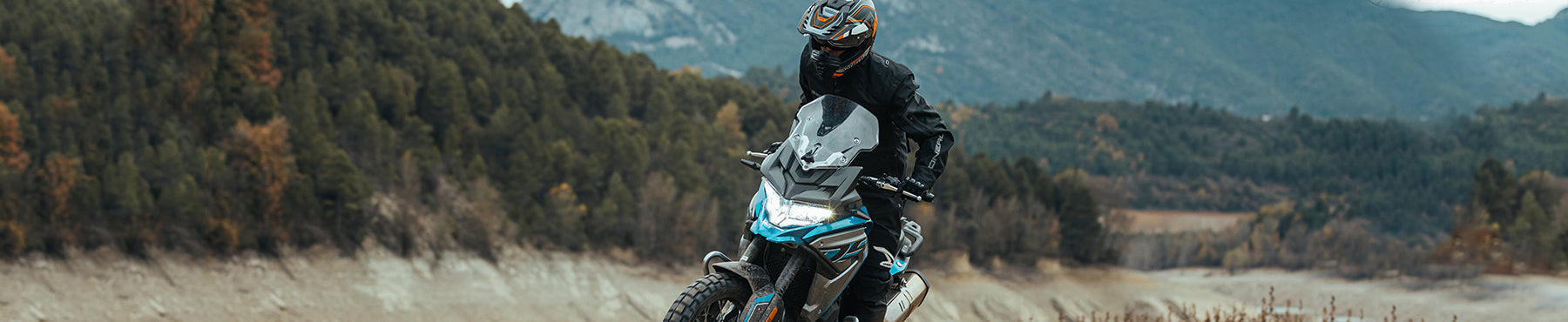 Eine Person in Schutzkleidung fährt mit einem Motorrad im Gelände, umgeben von einer Landschaft aus Bäumen und Bergen unter einem bewölkten Himmel.