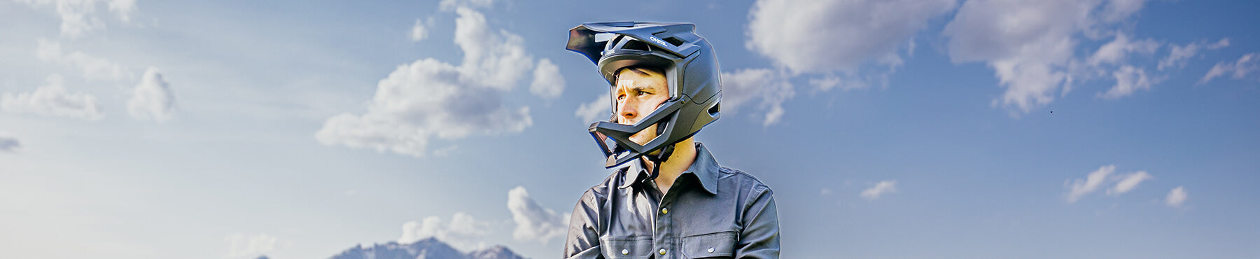 Eine Person mit Integralhelm blickt zur Seite unter einem blauen Himmel mit einigen Wolken. Im Hintergrund sind schwach Berge zu erkennen.