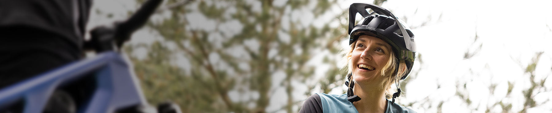 Eine Person mit Helm lächelt und blickt auf eine Person außerhalb des Bildes, vor einem unscharfen natürlichen Hintergrund. Sie trägt Outdoor-Radsportkleidung.