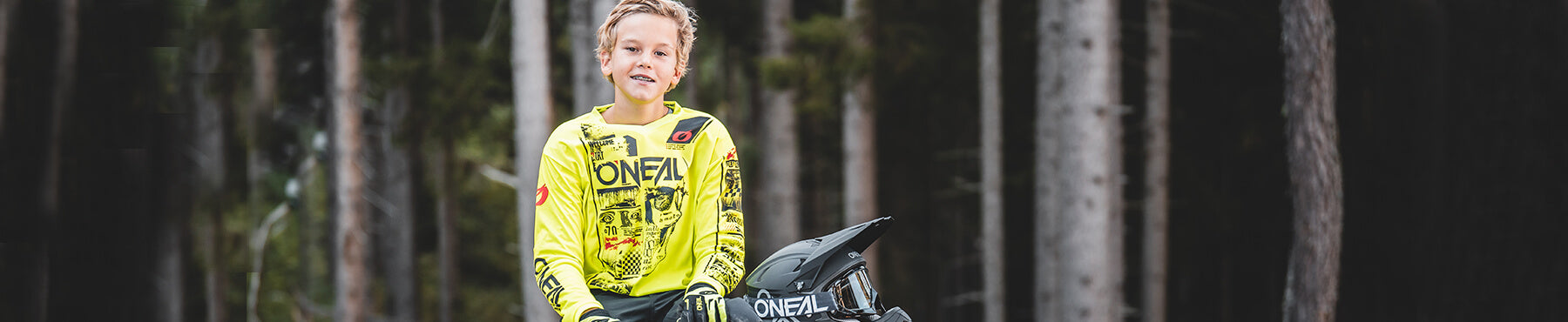 Ein junger Mensch in einem leuchtend gelben Radtrikot sitzt auf einem Geländemotorrad in einer Waldlandschaft und trägt einen Helm auf dem Schoß. Im Hintergrund sind hohe Bäume und eine verschwommene Waldszene zu sehen.