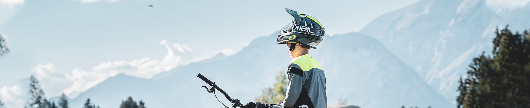Eine Person mit Helm und Schutzausrüstung steht mit einem Mountainbike da und blickt auf die Berglandschaft im Hintergrund. Der klare blaue Himmel und vereinzelte Wolken bereichern die Landschaft.