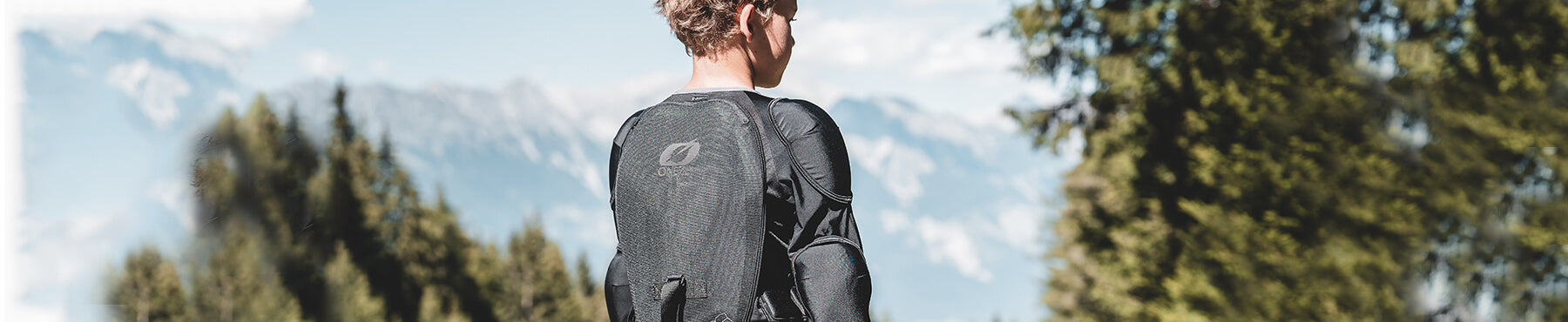 Eine Person in Schutzkleidung steht im Freien mit verschwommenen Bergen im Hintergrund. Der Fokus liegt auf ihrem Rücken, der einen netzartigen Anzug zeigt. Bäume rahmen die Szene unter einem klaren Himmel ein.