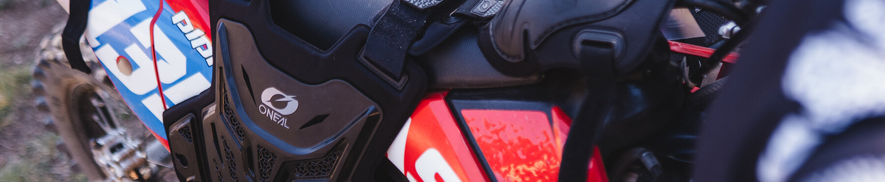 Nahaufnahme eines Motocross-Motorrads mit einem leuchtend roten und blauen Design. Auf dem Sitz des Motorrads ist Schutzausrüstung festgeschnallt, auf der das O'Neal-Logo zu sehen ist.