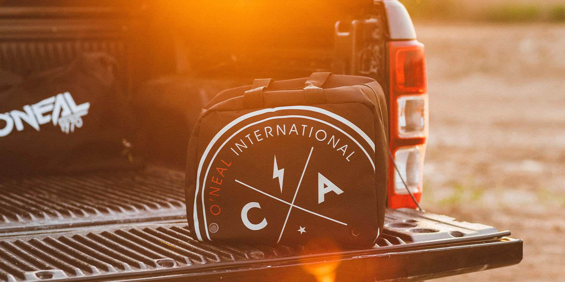 Eine schwarze Tasche mit einem runden „O'NEAL International“-Logo liegt auf der Ladeklappe eines Pickups. Die Sonne geht unter und wirft einen warmen Schein und Lichtreflexe über die Szene. Im Hintergrund der Ladefläche ist eine weitere Tasche zu sehen.