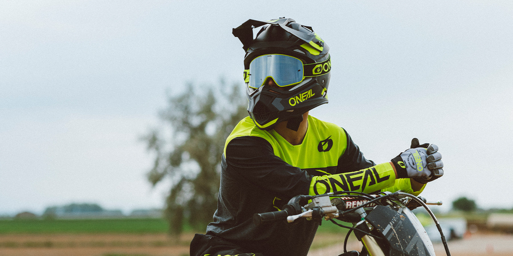 Ein Motocrossfahrer in leuchtend gelber und schwarzer Ausrüstung, einschließlich Helm, Schutzbrille, Handschuhen und Oberteil, sitzt auf einem Dirtbike. Im Hintergrund ist eine verschwommene ländliche Landschaft mit Feldern und einem Baum unter einem bewölkten Himmel zu sehen. Auf der Ausrüstung des Fahrers ist der Markenname „O'Neal“ zu sehen.