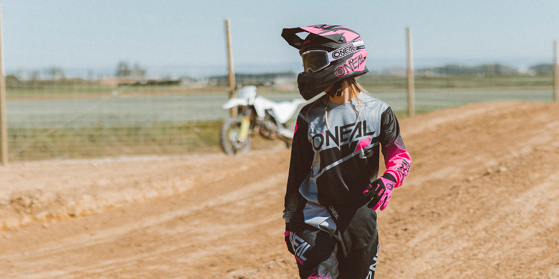 Eine Person in einem rosa-schwarzen Motocross-Outfit und Helm steht auf einer unbefestigten Strecke, im Hintergrund parkt ein weißes Dirtbike. Die Szene scheint im Freien an einem sonnigen Tag mit klarem Himmel zu spielen.