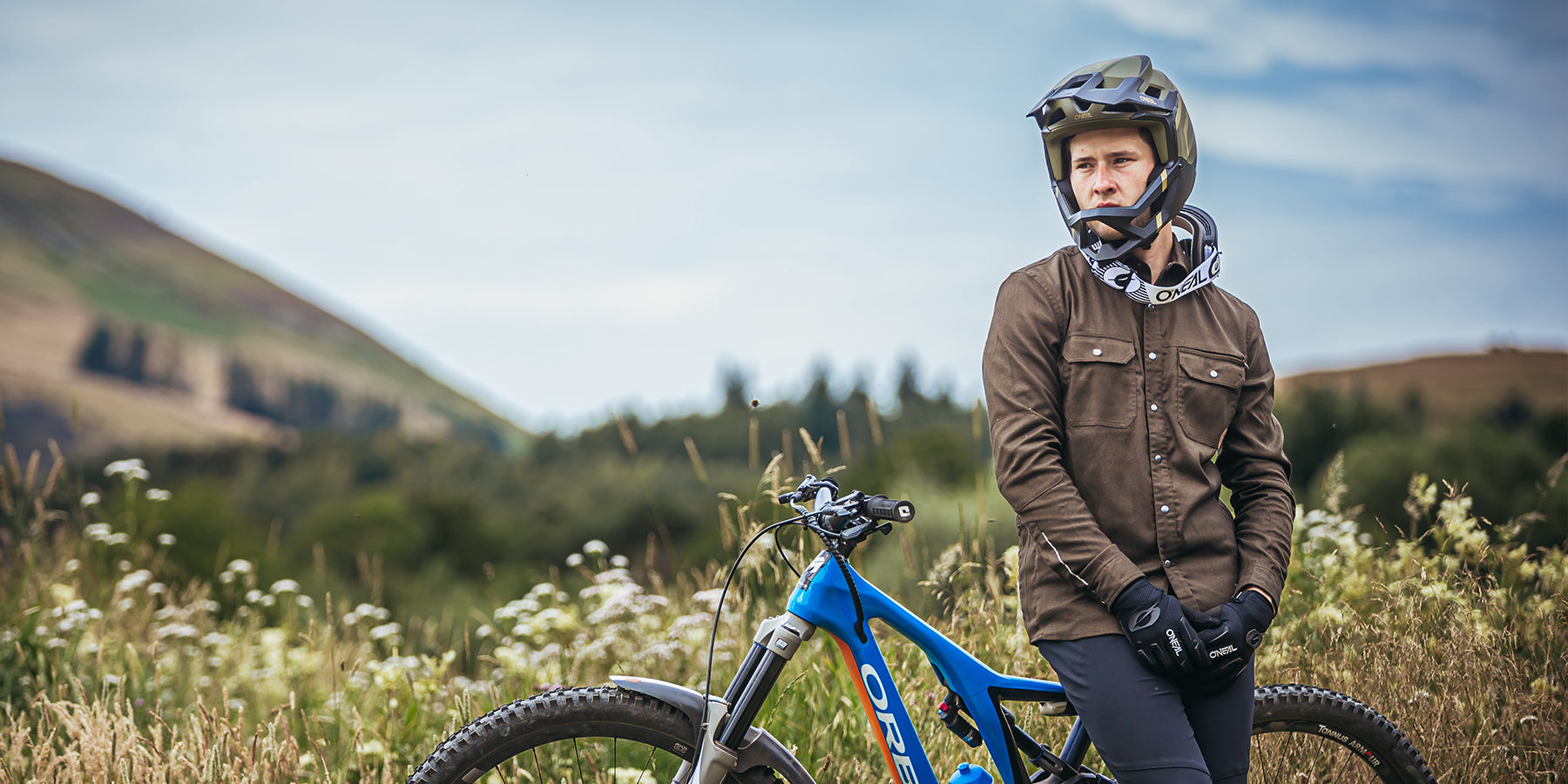 Eine Person mit Helm und Handschuhen lehnt an einem blauen Mountainbike auf einer Wiese mit sanften Hügeln und einem bewölkten Himmel im Hintergrund. Die Person trägt eine braune Jacke und schwarze Hosen und blickt in die Ferne.