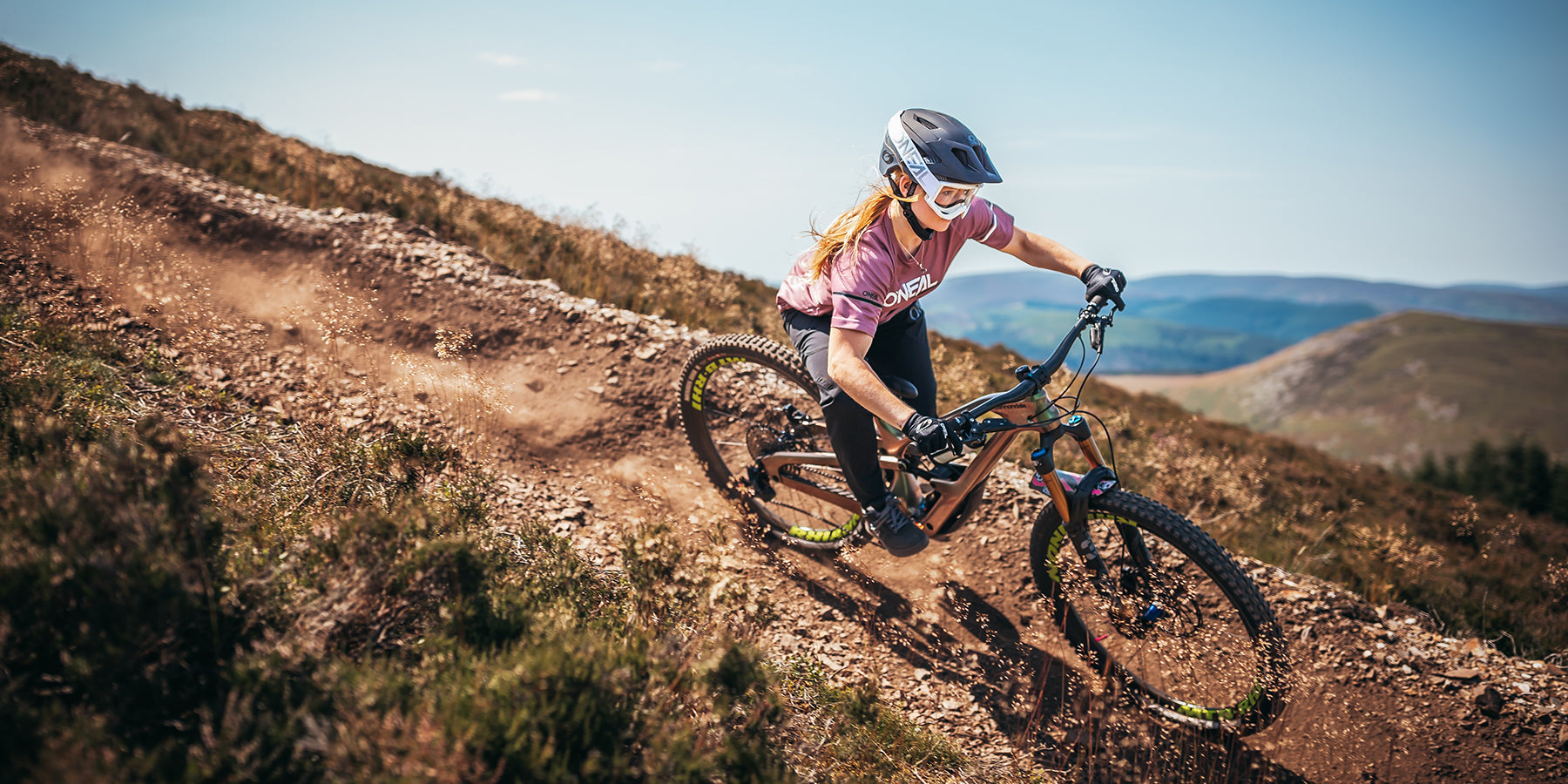 Eine Person mit Helm und Schutzausrüstung fährt an einem sonnigen Tag mit einem Mountainbike einen felsigen Pfad hinunter. Der Fahrer legt sich in eine Kurve und wirbelt dabei Staub und kleine Steine auf. Im Hintergrund sind sanfte Hügel und ein klarer blauer Himmel zu sehen.