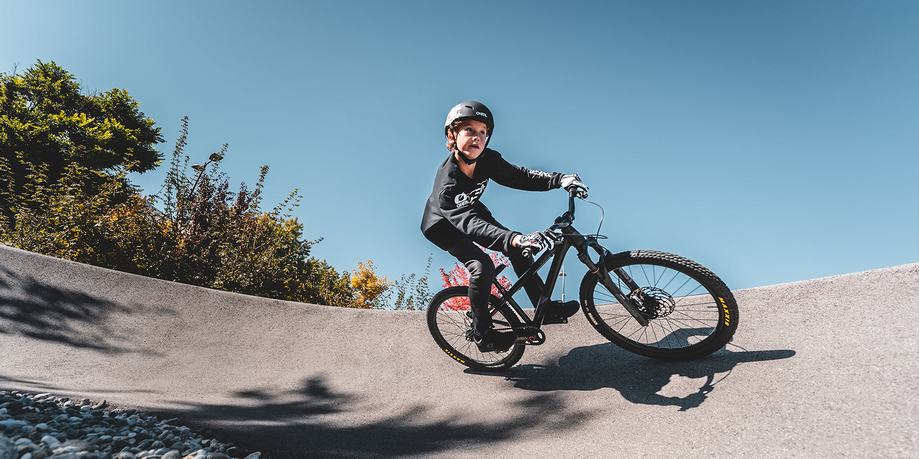 Ein Kind mit Helm und Radsportausrüstung fährt mit einem Mountainbike auf einer kurvigen Betonstrecke unter einem klaren blauen Himmel. Im Hintergrund säumen Bäume mit grünem und rotem Laub die Strecke. Das Kind scheint beim Navigieren einer Kurve vorsichtig das Gleichgewicht zu halten.