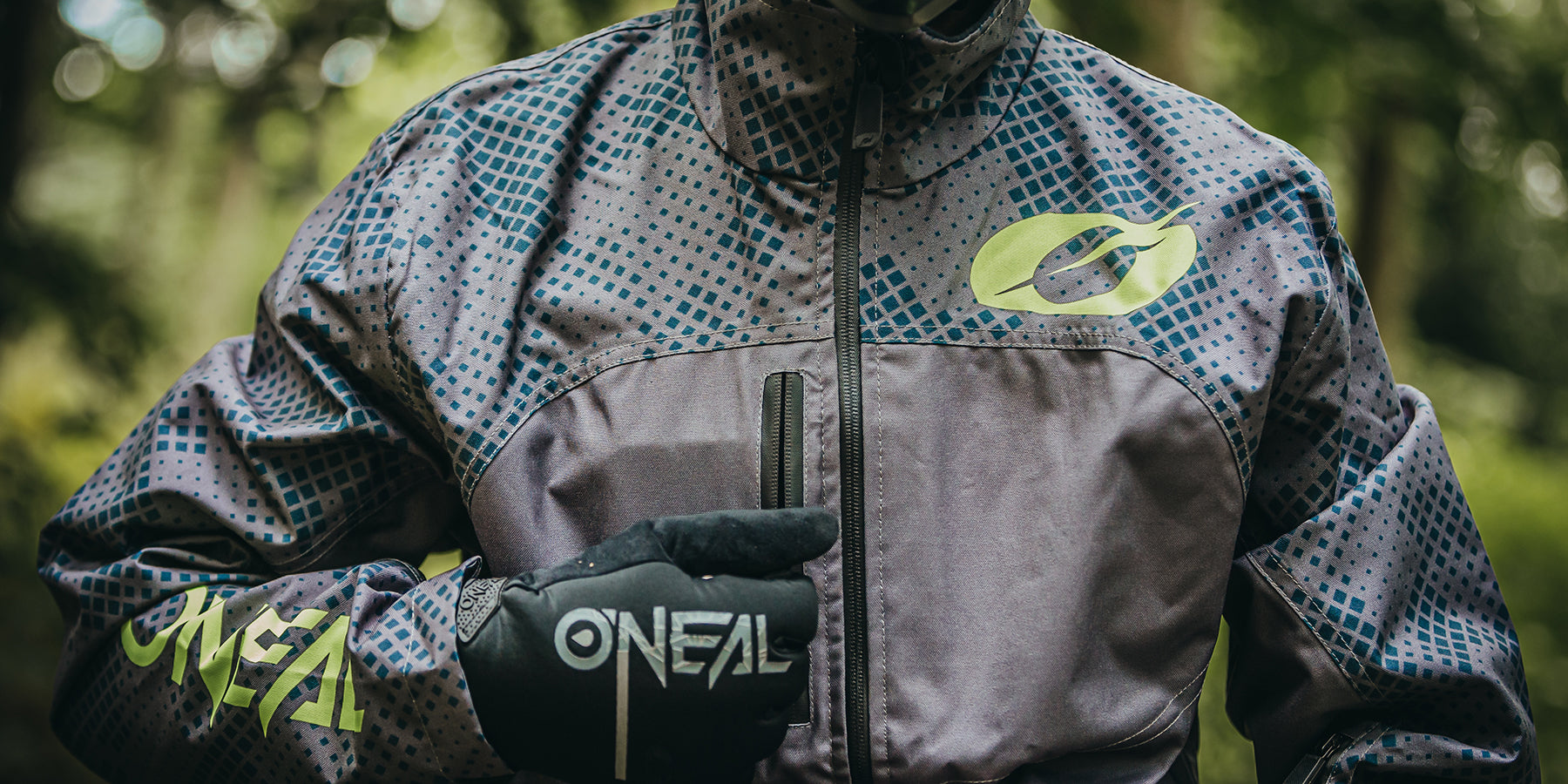 Nahaufnahme einer Person, die eine dunkle, gemusterte Jacke und Handschuhe mit der Marke „O'Neal“ trägt. Die Jacke hat einen Reißverschluss und grüne Akzente auf Brust und Arm. Die Person befindet sich in einer Außenumgebung mit sichtbarem Grün im Hintergrund.