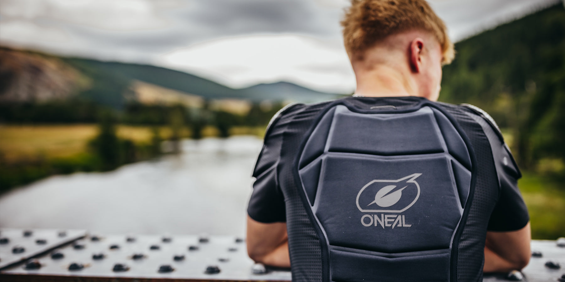 Eine Person mit kurzen blonden Haaren trägt eine schwarze O'Neal-Schutzweste und steht auf einer Brücke mit Blick auf eine malerische Landschaft mit einem Fluss, Bäumen und Hügeln im Hintergrund unter einem bewölkten Himmel. Die Person blickt von der Kamera weg.