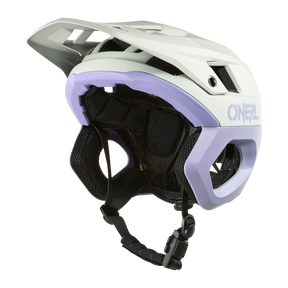 Ein weißer und lavendelfarbener O'NEAL TRAPPER Helmet SOLID Mountainbike-Helm mit Visier, seitlichen Belüftungsöffnungen, schwarzer Polsterung und einem verstellbaren Kinnriemen wird vor einem schlichten schwarzen Hintergrund gezeigt.