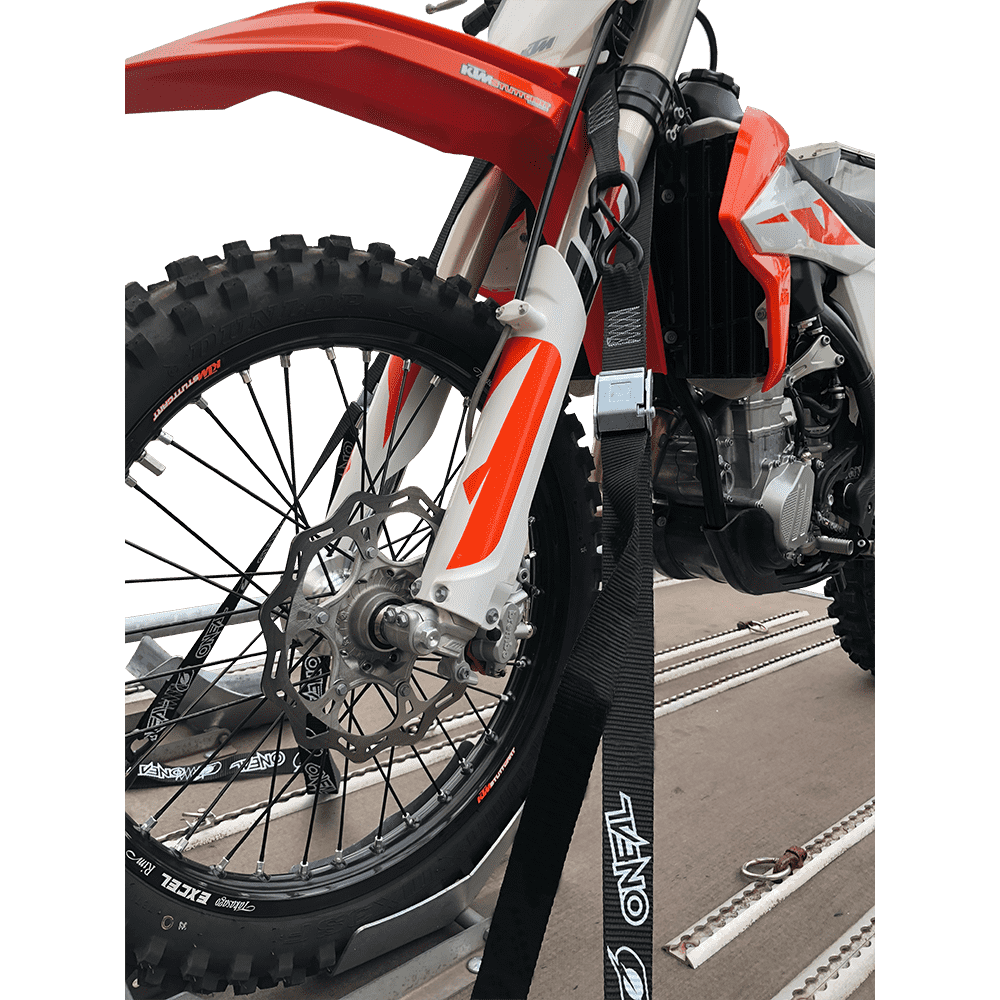 Nahaufnahme eines rot-weißen Geländemotorrads, das auf einem Anhänger mit O'NEAL DE LUXE Zurrgurte LOGO an der Vordergabel befestigt ist. Die Riemen der Marke O'NEAL halten das Fahrrad, wobei das Vorderrad und die Federung deutlich zu sehen sind.